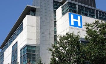 A modern, multi-story hospital building with windows and a prominent blue sign with a white 